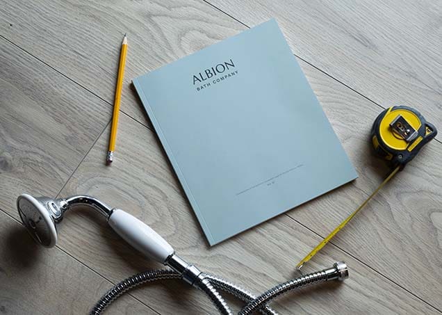 A4 blue Albion Bath Co Ltd brochure with measuring tape, pencil, and bathroom fittings on light wooden floor, showcasing Albion Bath Co's premium bathroom products.