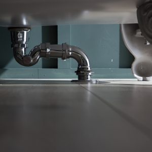 Exposed plumbing under bathroom sink with chrome pipe and grey tile flooring.