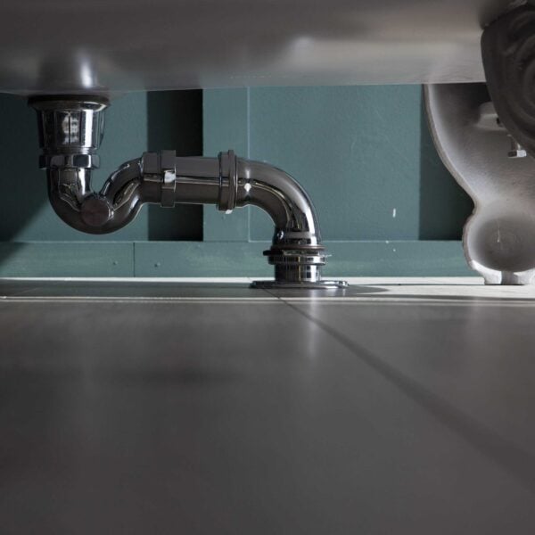Exposed plumbing under bathroom sink with chrome pipe and grey tile flooring.